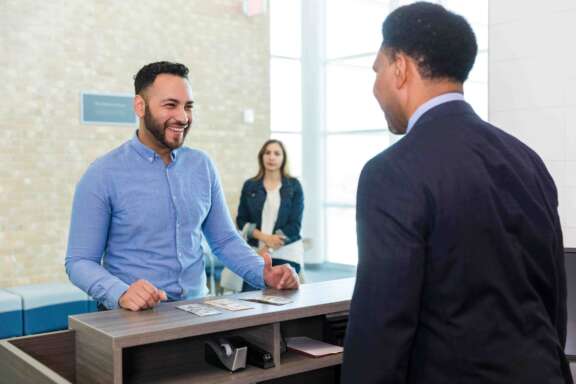Two cheerful men converse at a reception desk; one smiles broadly.