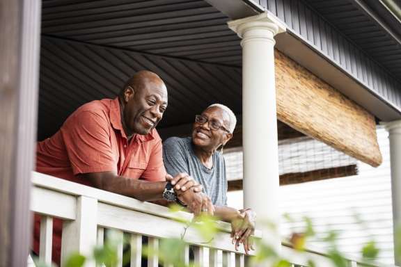 Three-quarter front view of relaxed Black seniors leaning on railing of restored home in Rockaway Beach, Queens, talking and smiling.