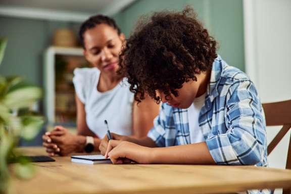 African American teen boy doing homework, making notes in the notebook at the table, side view.